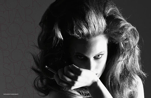 Monochrome portrait of a woman with voluminous hair, holding a cigarette to her mouth, against a blurred background with repeated text.