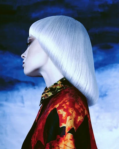 Side profile of a woman with platinum blonde bob haircut wearing a patterned top against a blue, cloud-like background. Fashion beauty.