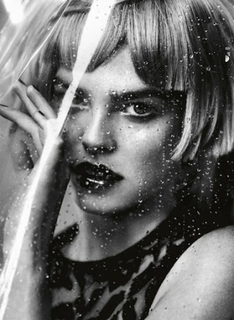 Monochrome portrait of a woman with short blonde hair looking through a glass covered in water droplets in a beauty shot.