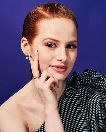 Close-up portrait of an adult female with red hair in black and white patterned dress, with light skin and purple background.