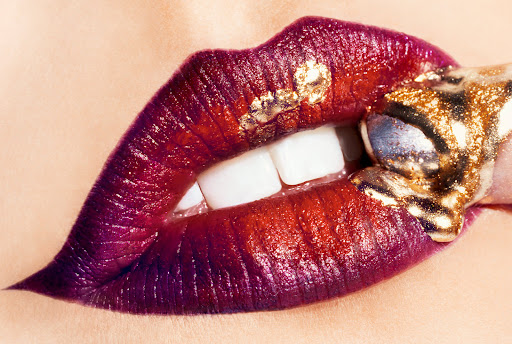 Close-up of a woman's red and purple lips with white teeth, biting a gold covered object, high-fashion beauty shot.