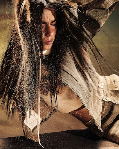 Woman with long hair poses while sand falls over her head and face, wearing a denim shirt in a studio setting, medium shot.