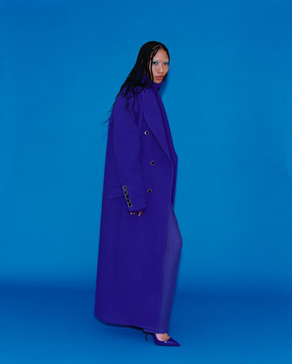 A woman in a purple double-breasted overcoat and matching purple pants and high heels strikes a fashion pose on a blue backdrop.