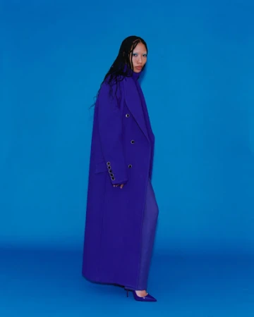 A woman in a purple double-breasted overcoat and matching purple pants and high heels strikes a fashion pose on a blue backdrop.
