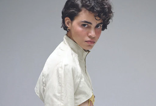 Portrait of a model with short dark curly hair wearing a light-colored high-collar jacket against a plain grey background