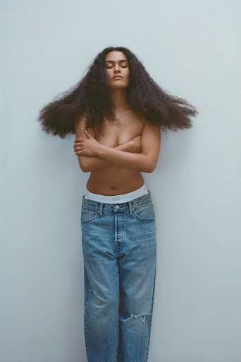 Woman with long dark curly hair standing against a plain wall wearing blue denim jeans and crossing arms over her chest