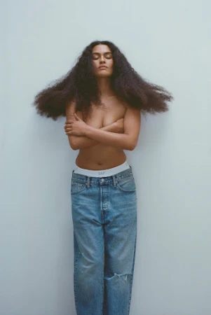 Woman with long dark curly hair standing against a plain wall wearing blue denim jeans and crossing arms over her chest