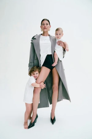 Woman holding an infant and hugging a toddler while wearing a grey trench coat and black heels against a white background