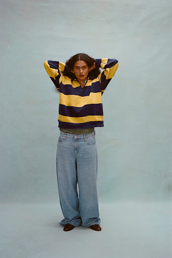 A woman stands in a photo studio wearing a striped yellow and navy long-sleeved shirt, light-wash denim jeans, and brown shoes.