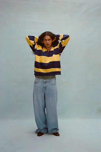 A woman stands in a photo studio wearing a striped yellow and navy long-sleeved shirt, light-wash denim jeans, and brown shoes.