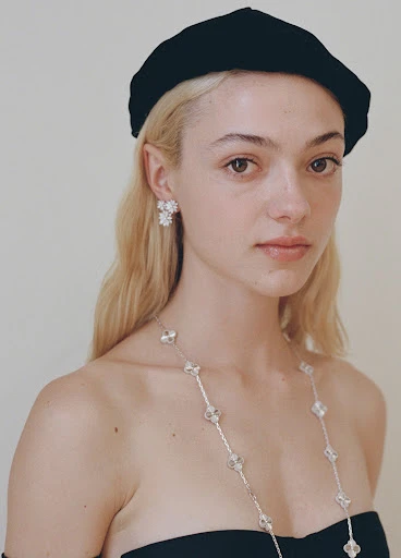 Close-up portrait of a fair-skinned woman with platinum blonde hair wearing a black beret, dress, earrings, and a long crystal necklace.