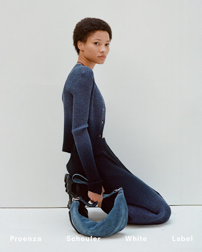 A woman with short dark hair models blue knitwear and a blue suede purse in a fashion studio against a plain backdrop.