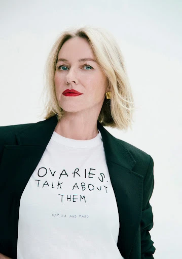 Close-up studio portrait of Naomi Watts with blonde hair, wearing a blazer and graphic tee shirt with red lipstick on light background.