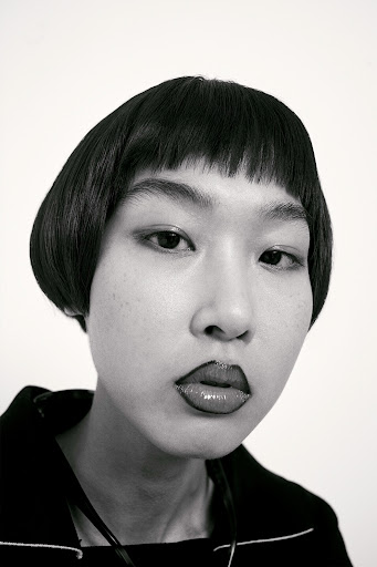 Close-up black and white portrait of a young woman with black bob haircut and bold lipstick against a plain white background.