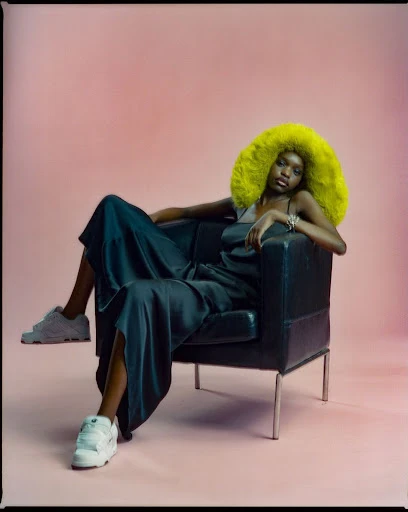 Fashionable Black woman with afro wearing a dress and sneakers, sitting in black chair against a pink gradient background in studio shot.