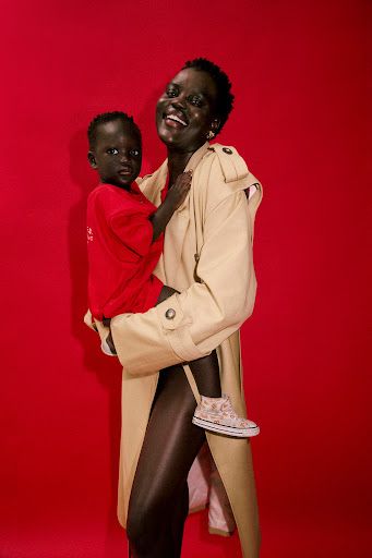 A dark-skinned woman holds a dark-skinned child in front of a red background. The woman wears a tan trench coat, and the child wears a red sweatshirt.