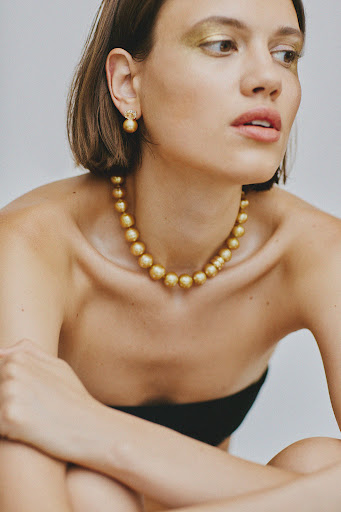 Portrait of a woman with bobbed hair wearing a golden necklace and earrings against a neutral backdrop. Gold makeup on eyelids.