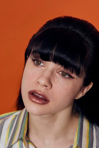 Portrait of a woman with dark hair and a striped shirt against an orange background, looking off-camera with neutral facial expression.
