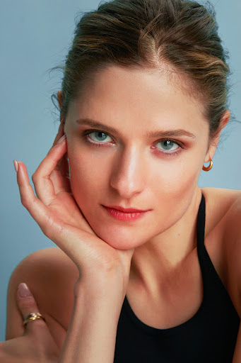 Close-up portrait of a woman with her hand on her chin, wearing a black top against a plain grey-blue studio backdrop.