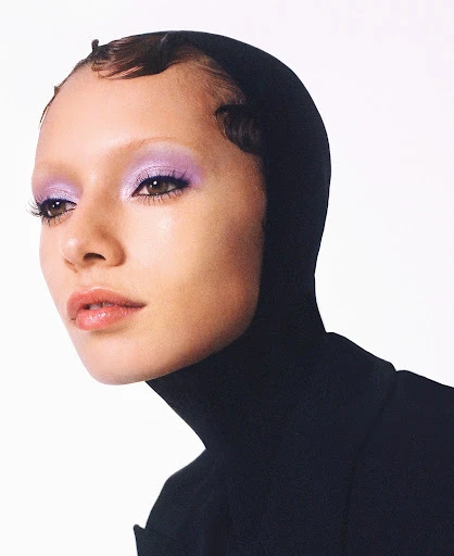 Headshot of a woman with purple eyeshadow and a black turtleneck sweater, posing in front of a white background, looking off to the side.