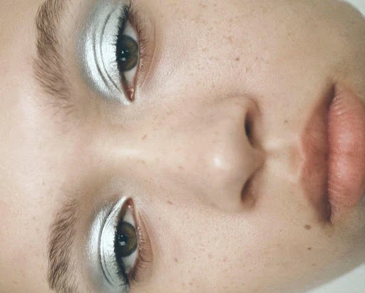 Close-up studio shot of a young woman with light skin and striking silver eyeshadow. Features focus on the eyes, nose and mouth.