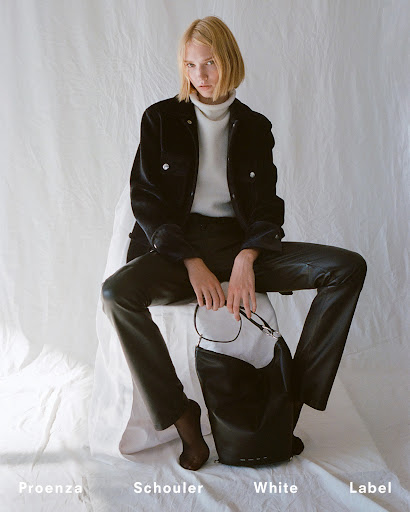 Fashion shoot of a single model wearing a black jacket and black pants, seated with a black and white bag in a light studio setting.