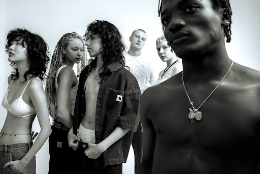 Black-and-white studio shot of six models in diverse styles, including a shirtless man with a necklace, against a bright white backdrop.