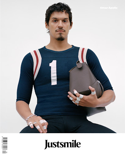 Portrait of a person with a football jersey holding a brown bag, wearing multiple rings on each finger, studio lit on a white background.