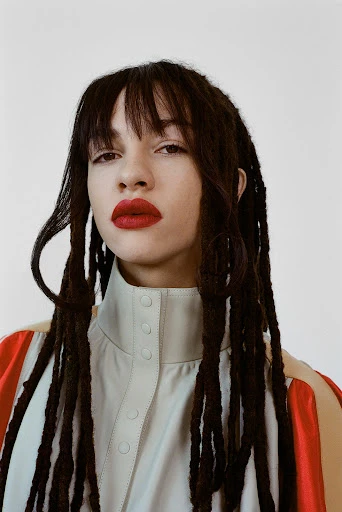 Portrait of a woman with dreadlocks and bangs, wearing a light-colored collared shirt and red lipstick, against a white background.