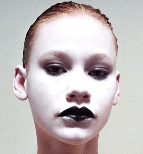 Close-up portrait of a young person with white face paint and dark lipstick, showcasing a striking and artistic beauty look in a neutral indoor setting.
