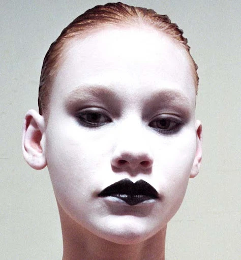 Close-up portrait of a young person with white face paint and dark lipstick, showcasing a striking and artistic beauty look in a neutral indoor setting.