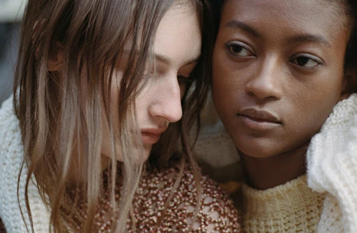 Two young women pose together, one with fair skin and the other with dark skin, both wrapped in a cream-colored knit textile.