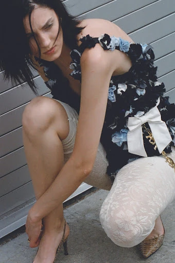 Young woman in black floral dress with white lace tights and snake print heels poses against a gray corrugated metal background.