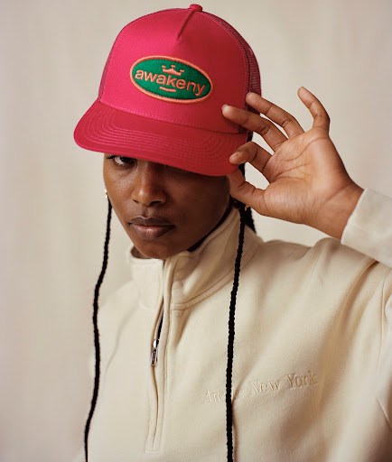 Close-up of a Black woman wearing a pink cap and off-white half-zip sweatshirt, looking directly at the camera against a neutral background.