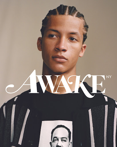 Portrait of a young adult male with cornrow braids. He wears a black graphic T-shirt and striped knit sweater against a beige background.