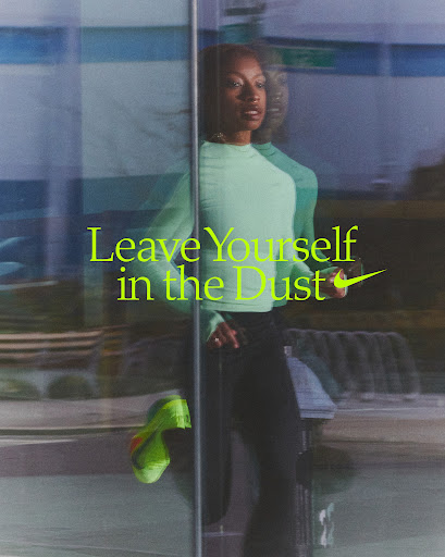 A woman in athletic apparel stands behind glass with a neon ad for Nike running shoes, styled with green top, black pants and green shoes.