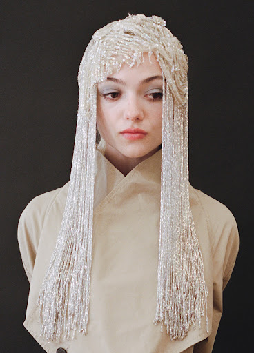 Young woman with neutral expression wearing tan trench coat and elaborate beaded headpiece with long, flowing beaded fringe over dark background.