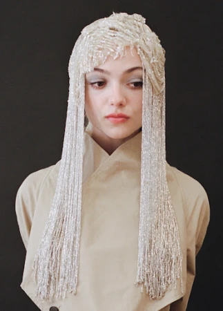 Young woman with neutral expression wearing tan trench coat and elaborate beaded headpiece with long, flowing beaded fringe over dark background.