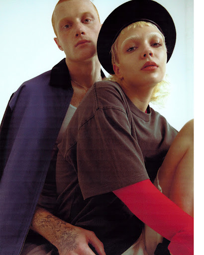 Two people pose in a studio. One with red hair and blue jacket stands behind, while the other in a black hat sits. 