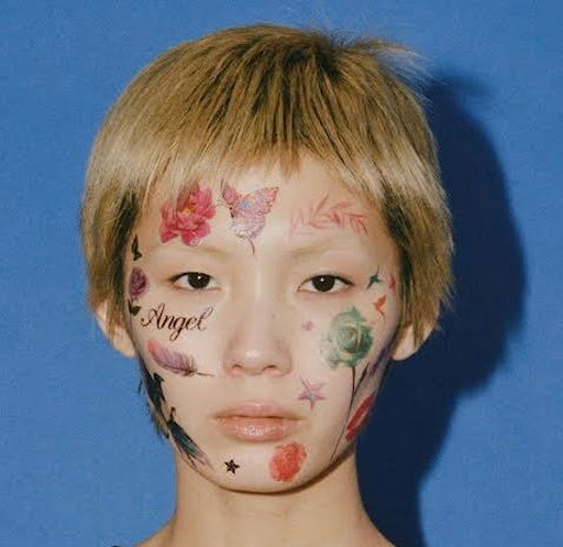 Close-up of a person with floral and feather face paint in red, pink, green, and black on a light skin tone against a blue background.