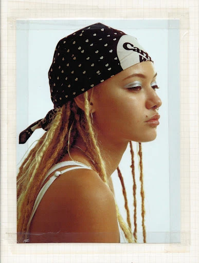 Portrait of a young woman with blonde dreadlocks, wearing a black and white headscarf, styled with silver eyeshadow and lip gloss.