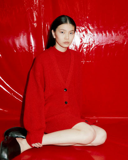 Asian woman poses indoors in a red cardigan and black shoes against a vivid red backdrop, creating a bold, monochromatic fashion shot.