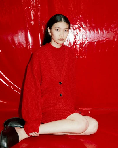 Asian woman poses indoors in a red cardigan and black shoes against a vivid red backdrop, creating a bold, monochromatic fashion shot.