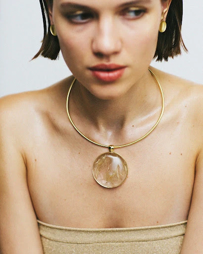 Portrait of a young woman with short hair wearing a gold tube top, gold necklace, and gold earrings against a neutral background.