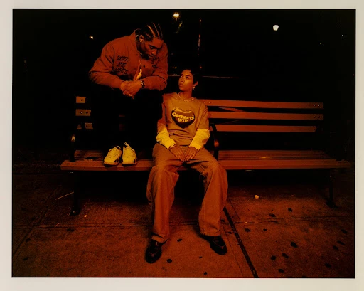 Two people, one standing and one sitting, on a bench at night. The person sitting is wearing a t-shirt with logo.
