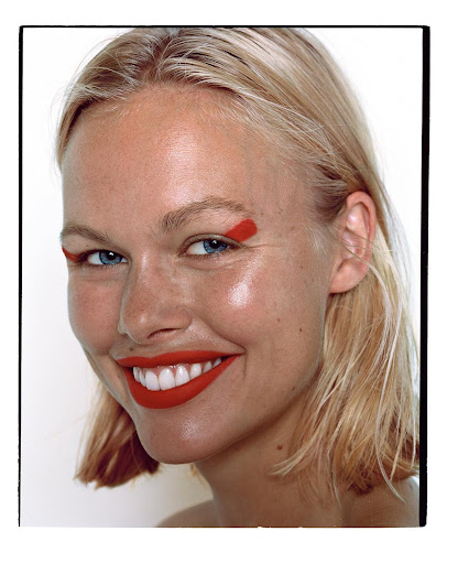 Close-up portrait of a smiling blonde woman with red lipstick and graphic red eye makeup against a plain white background.