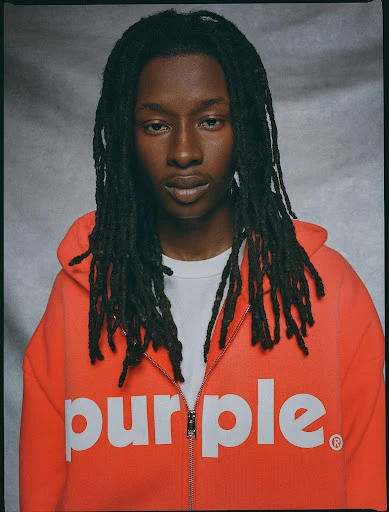 Portrait of an adult Black male with dreadlocks wearing an orange zip-up hoodie with 'purple' printed on the front, in a studio setting.