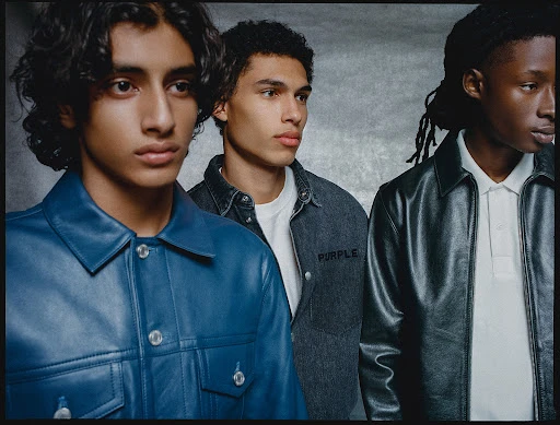 Three young men posing together wearing leather and denim jackets against a textured grey background.