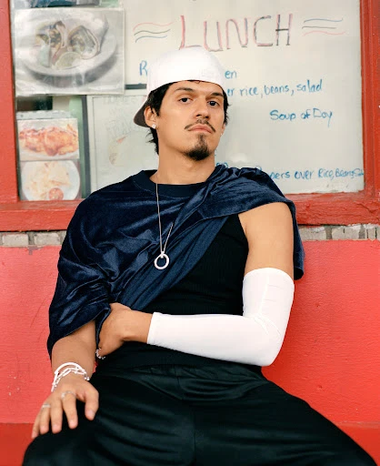 Young man poses on red bench in front of lunch menu, wearing cap, black top/pants, and draped blue velour fabric in an urban setting.