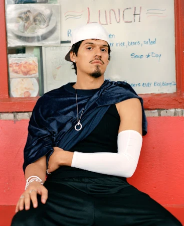 Young man poses on red bench in front of lunch menu, wearing cap, black top/pants, and draped blue velour fabric in an urban setting.